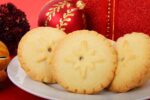 Three mince pies are arranged on a plate in front of a red Christmas present and bauble.